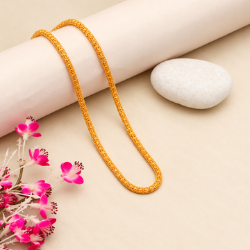 A gold chain necklace draped over a rolled cream-colored paper cylinder, with a smooth white stone and pink flowers on a light beige background.